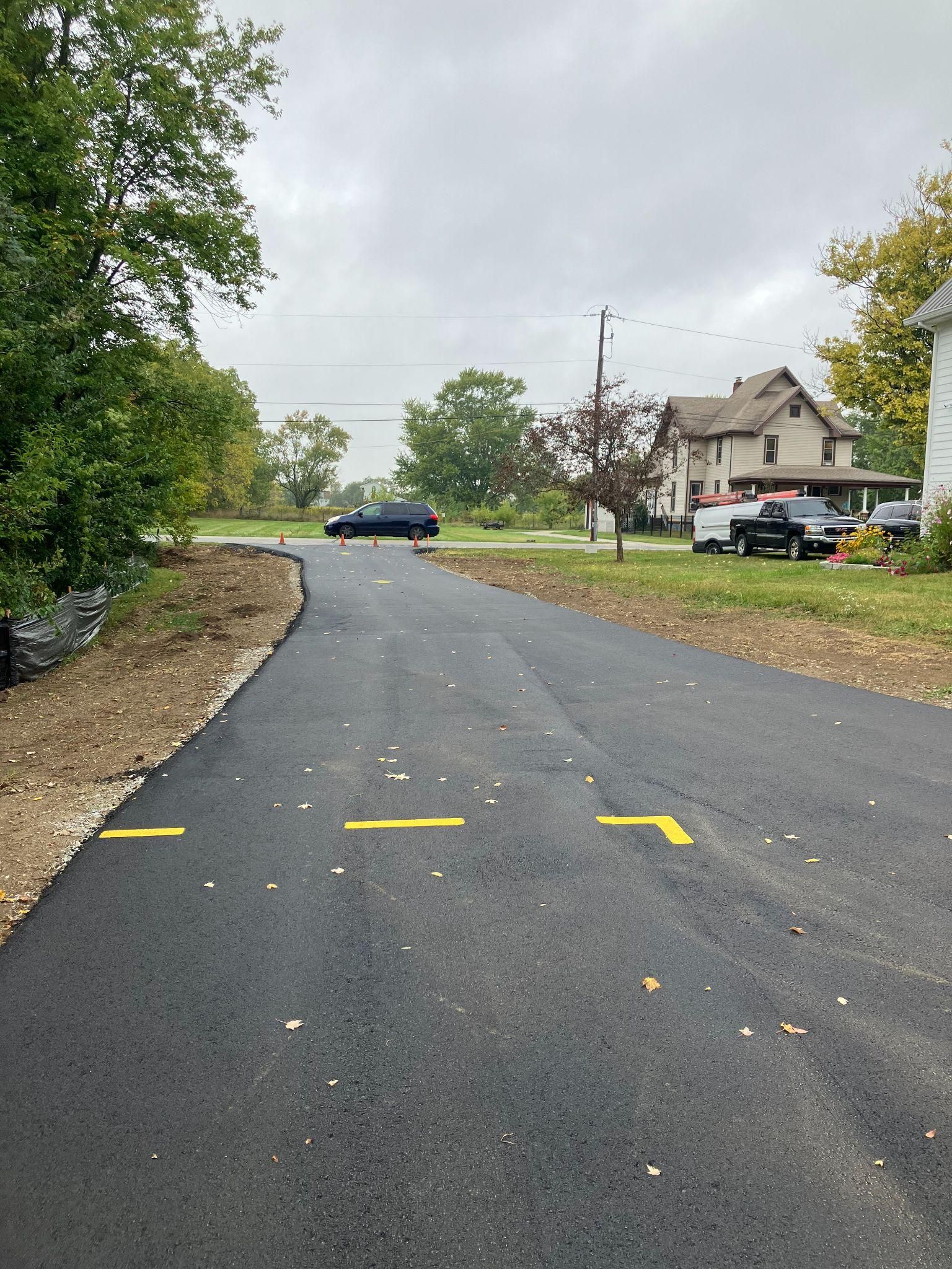 Newly paved asphalt road with yellow directional arrows, residential area, cloudy sky.
