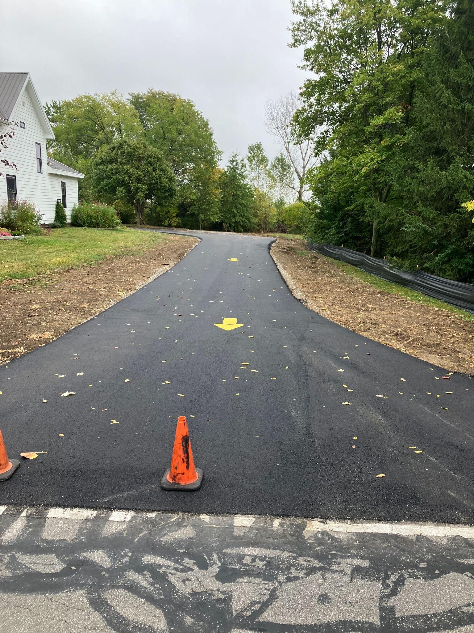 Asphalt driveway with orange cones and yellow directional arrows, leading to a house, under a cloudy sky.