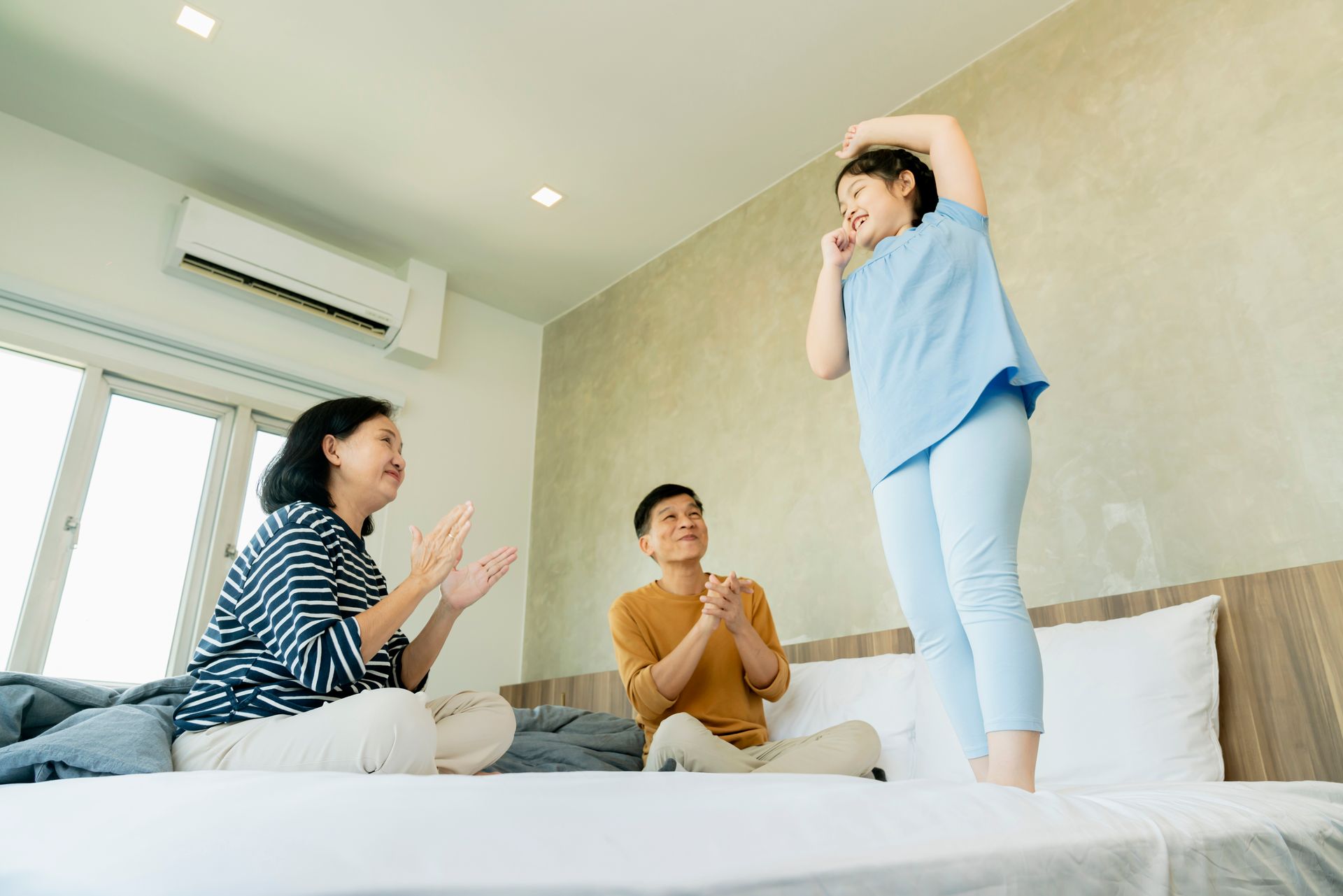 Three people on a bed in a bright bedroom, clapping while a child stands beside them with hands on head.