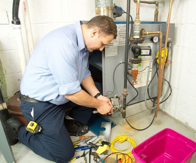 Technician kneeling beside a furnace, adjusting pipes and wiring in a utility room.
