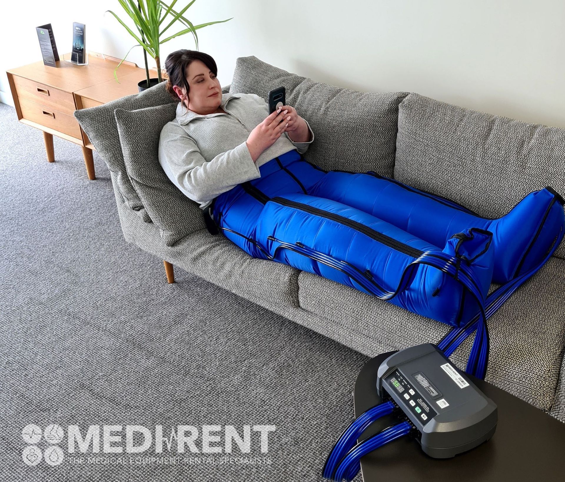 A person wearing blue pneumatic compression leg sleeves while resting on a couch and looking at a smartphone.