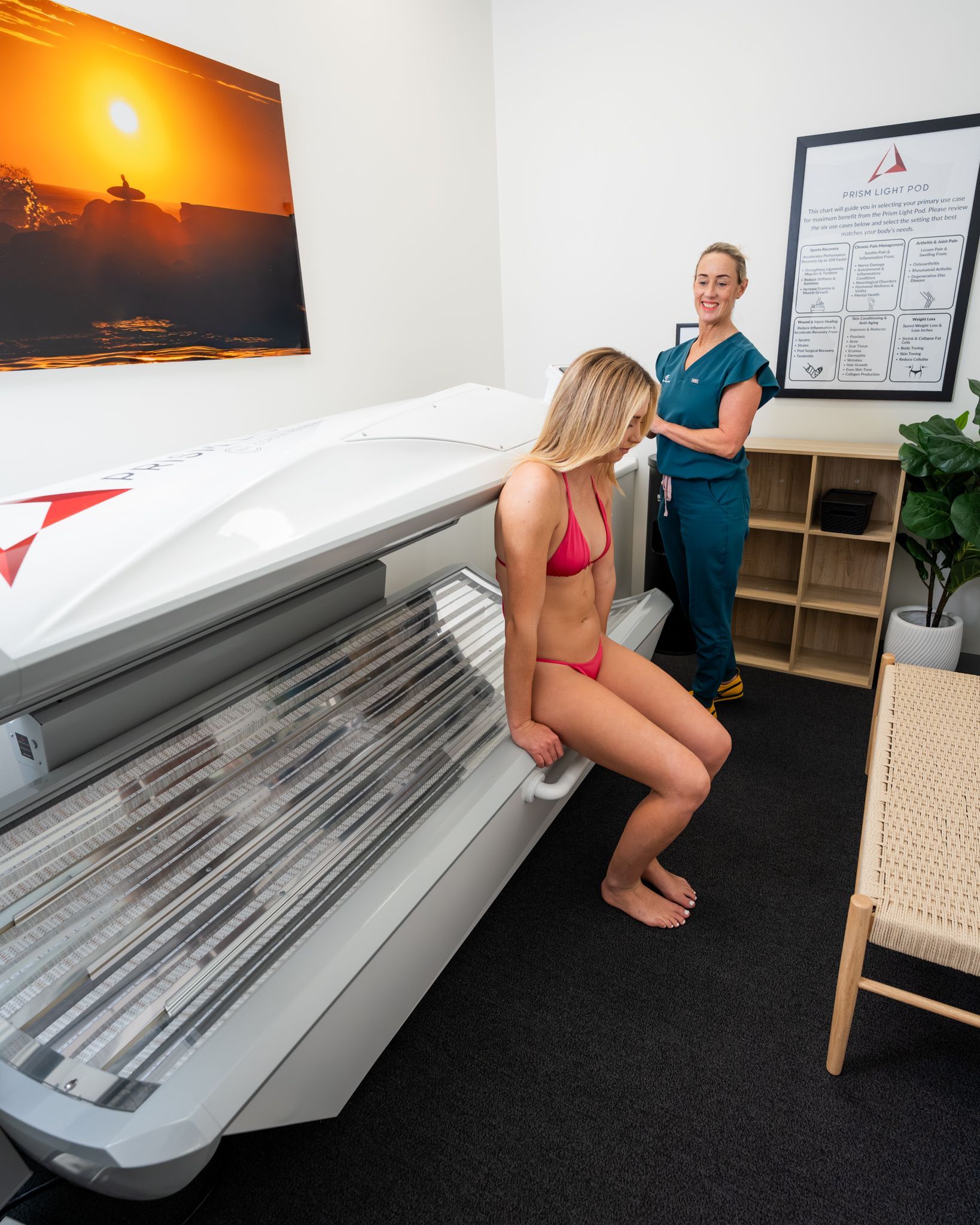 Woman in a bikini sits on a tanning bed as a smiling staff member watches. Room with decor.