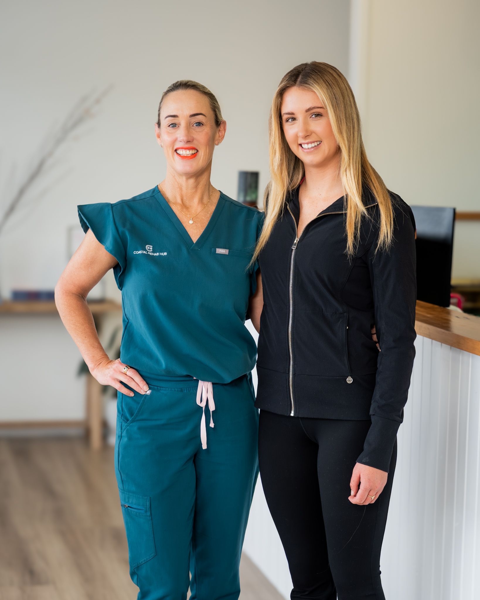 Three health professionals smiling, wearing teal scrubs, by a wooden reception desk.
