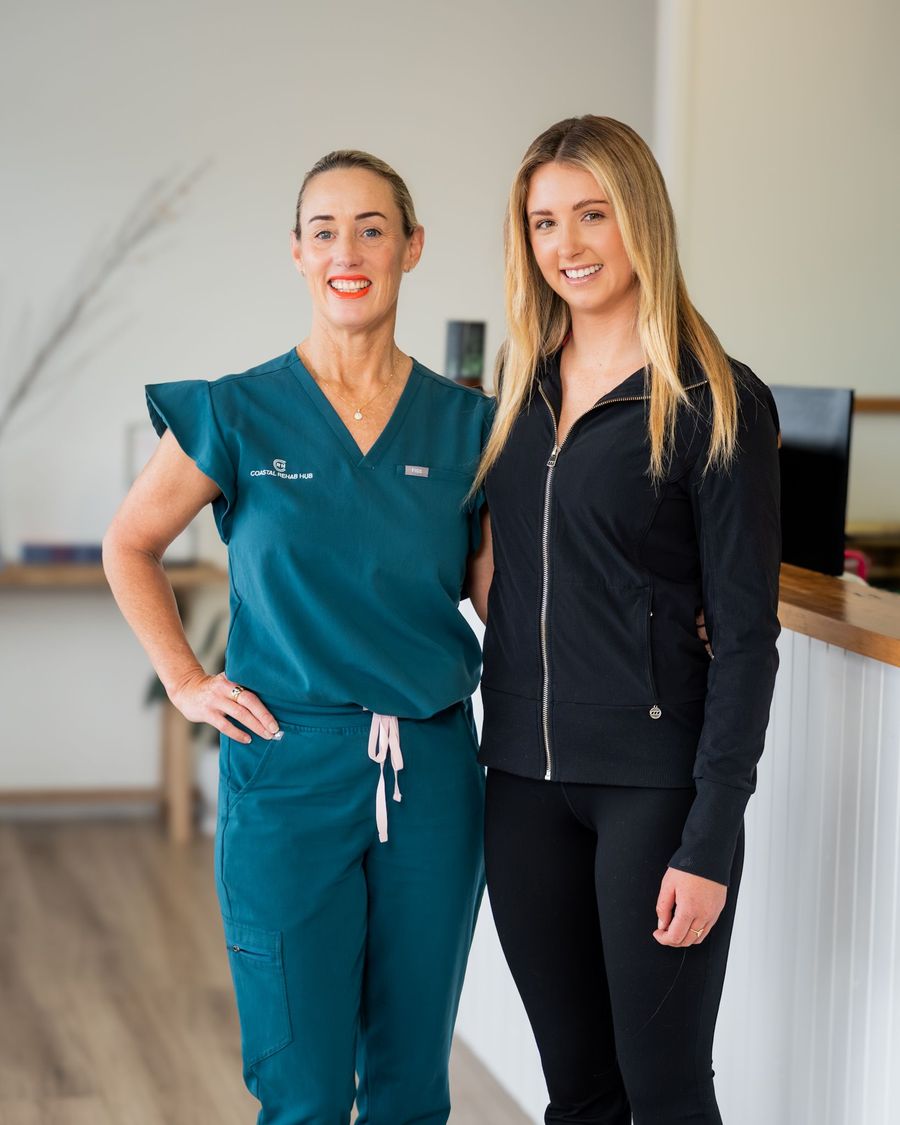 Two women smiling, one in teal scrubs, the other in a black jacket and pants, pose indoors near a reception desk.