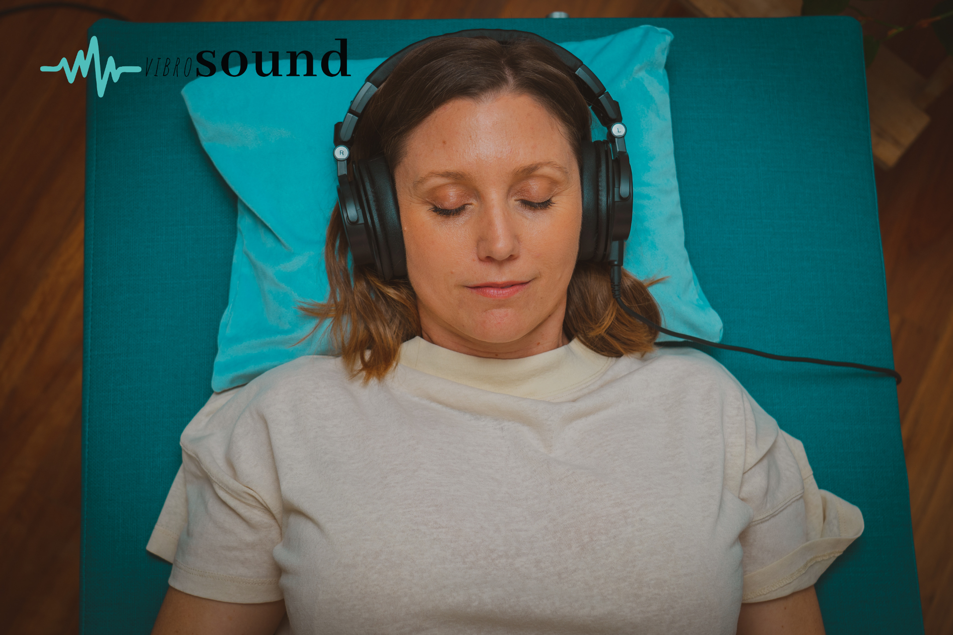 Man with headphones and closed eyes, holding a teal notebook, sitting in front of a bookcase.