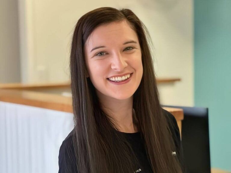 Woman with long dark hair smiles, in a bright office space.
