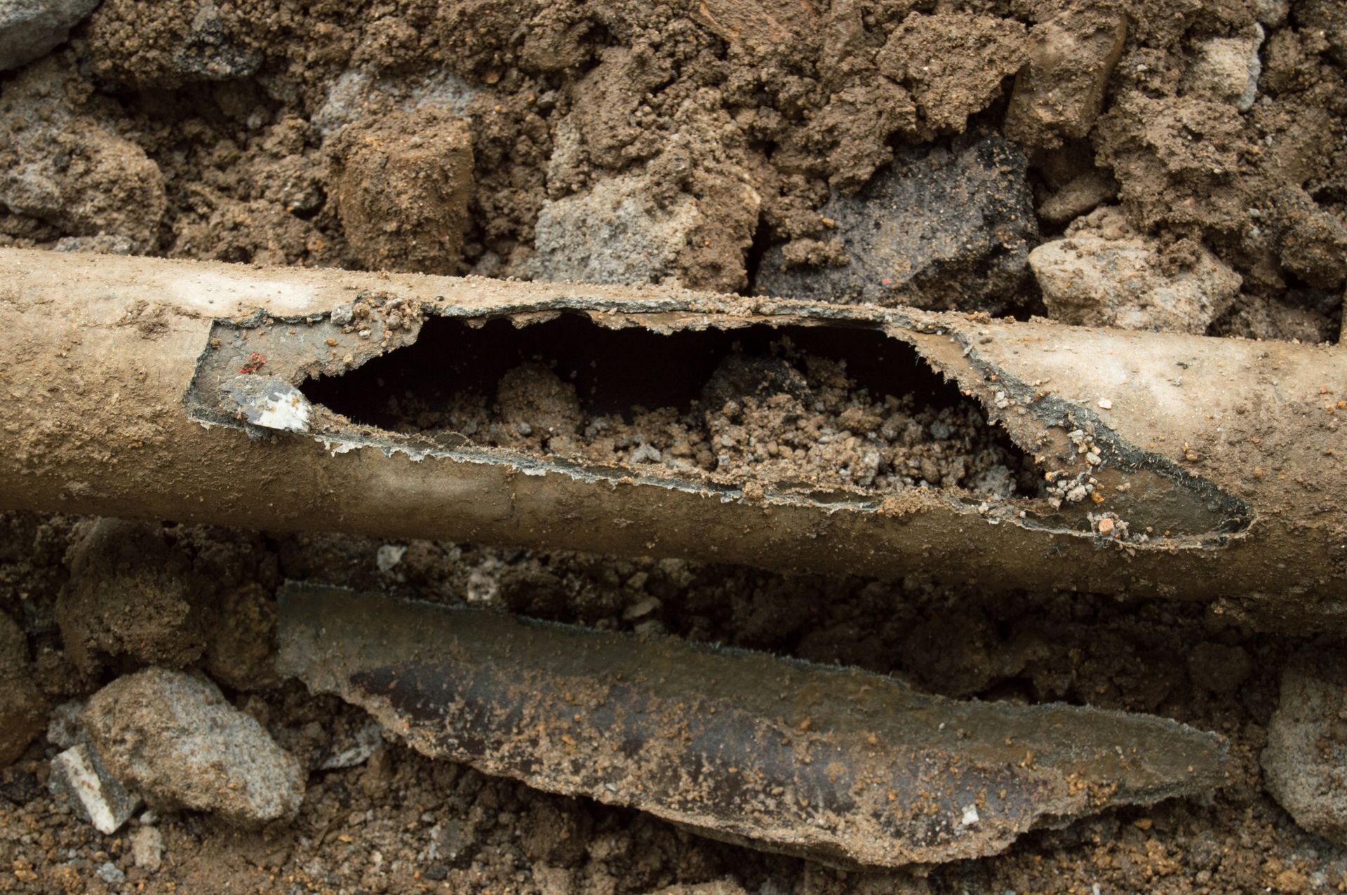 A corroded, broken pipe in soil, the interior filled with dirt, surrounded by loose earth