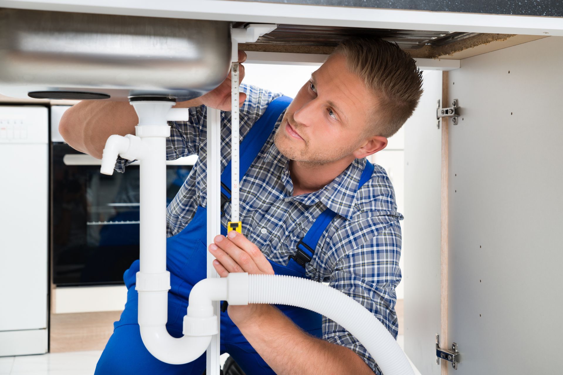 Plumber in blue overalls measuring kitchen sink pipes under counter for plumbing repair service.