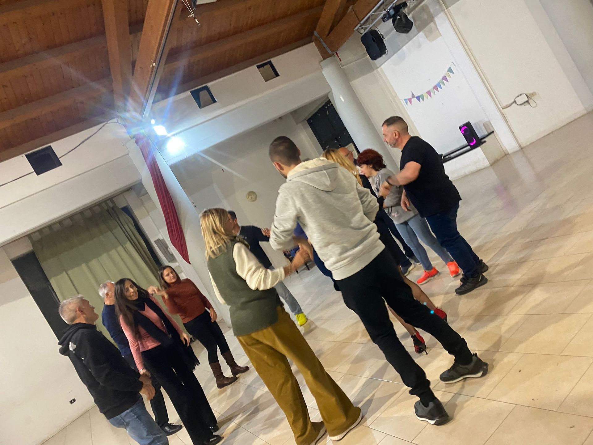 Persone che ballano in una sala da ballo illuminata. Alcuni si tengono per mano e provano i passi. Travi in ​​legno e pareti bianche.