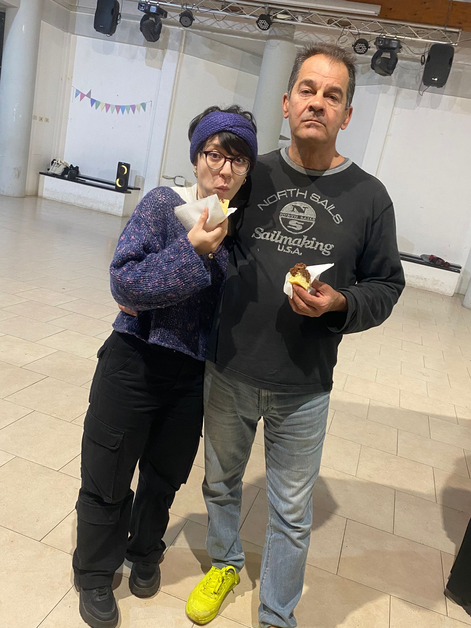 Due persone mangiano in una stanza. La donna indossa un maglione blu, pantaloni neri e una fascia per capelli. L'uomo indossa una camicia nera, jeans e scarpe gialle.