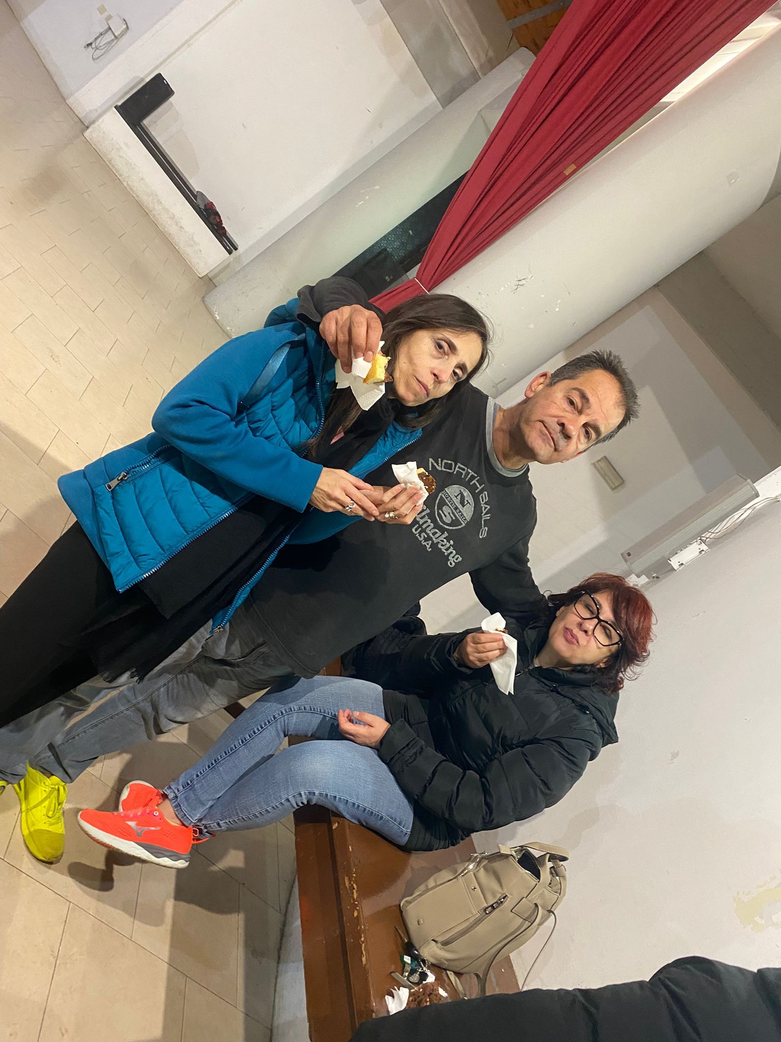 Tre persone in casa stanno mangiando: una con la giacca blu, un'altra con quella nera e una con le scarpe rosse e gli occhiali.