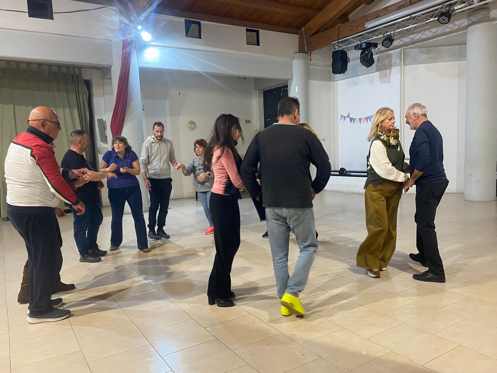 Persone che ballano in una sala illuminata. Le coppie si tengono per mano, mentre altri guardano.