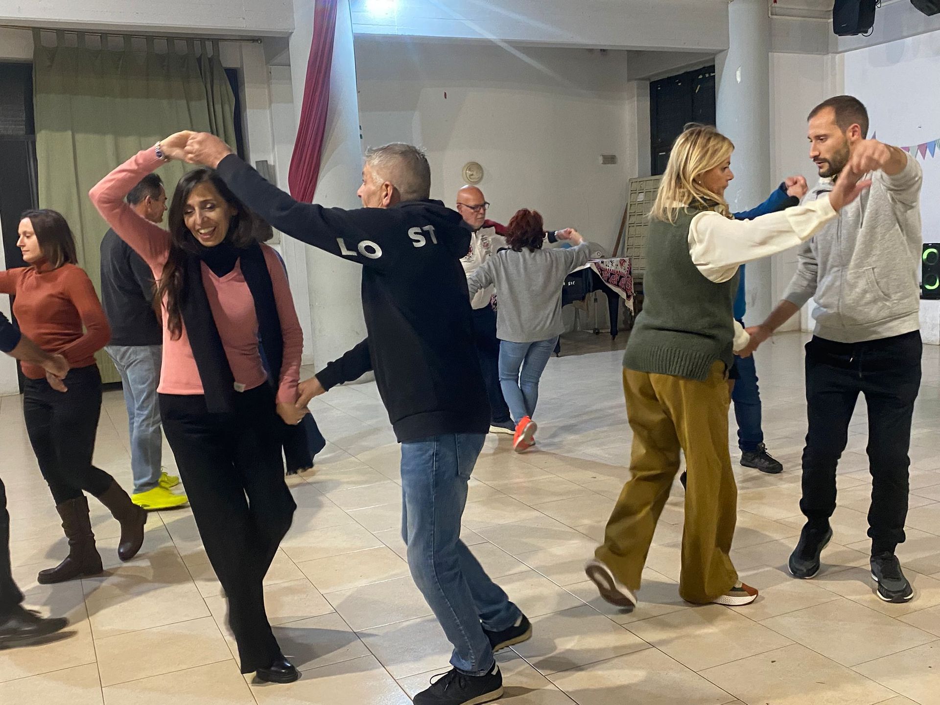 Persone che ballano in uno spazio interno ben illuminato, alcune in coppia, con le braccia alzate e sorridenti.