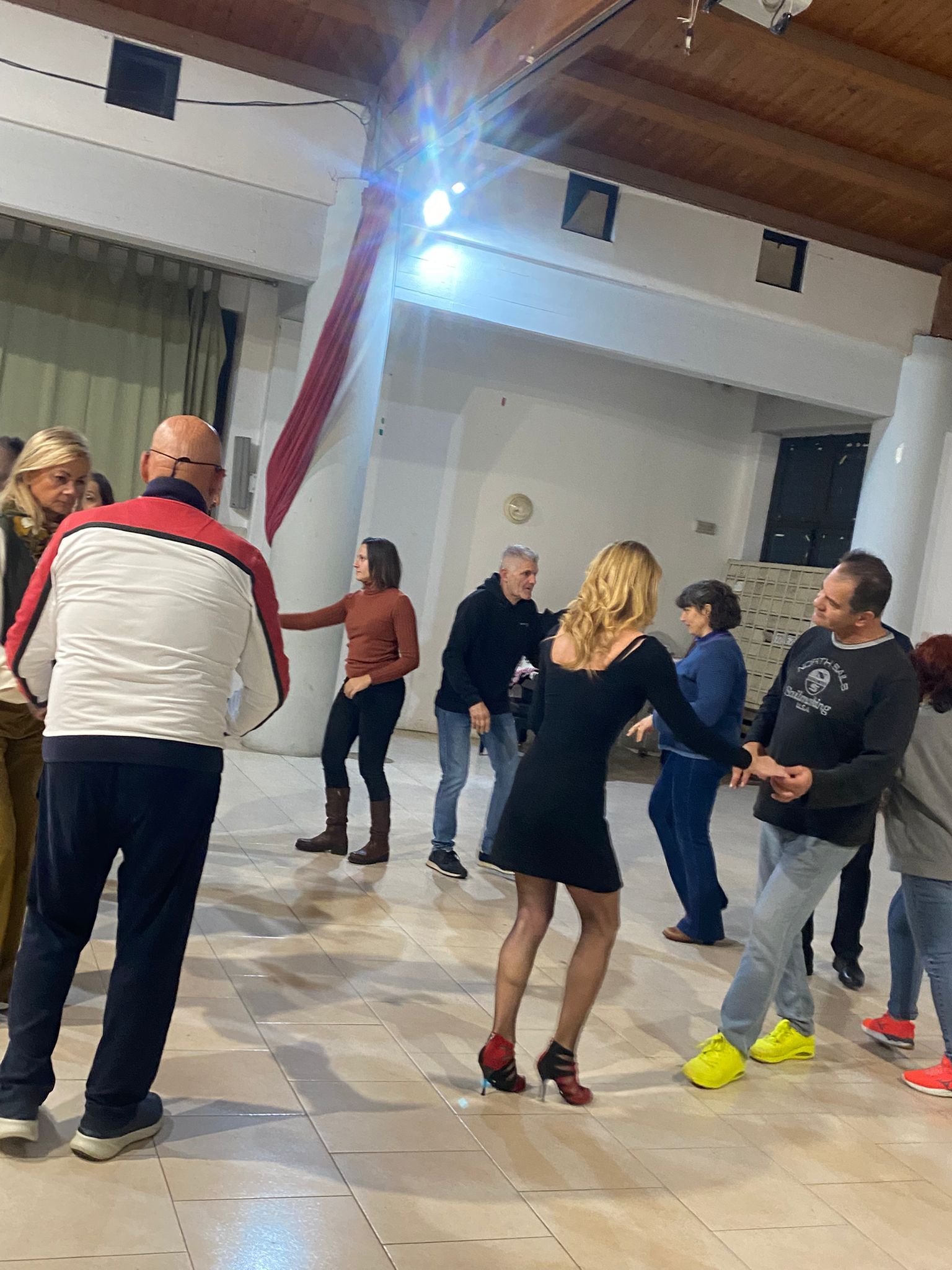 Persone che ballano in una stanza illuminata. Una donna in abito nero ne guida un'altra in una danza. Altri guardano e ballano.