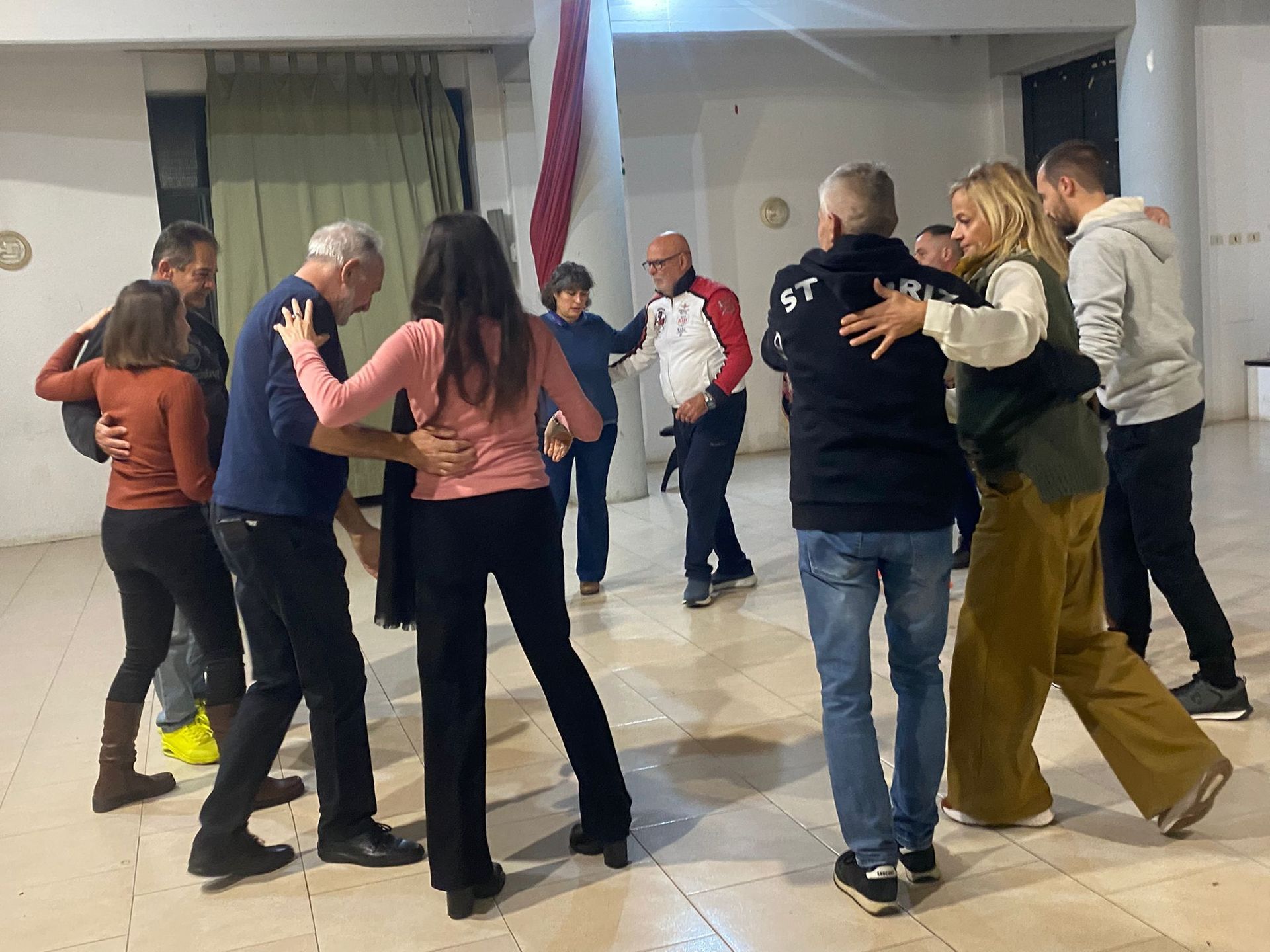 Persone che ballano in una stanza illuminata. Le coppie si abbracciano e si muovono, gli altri guardano.