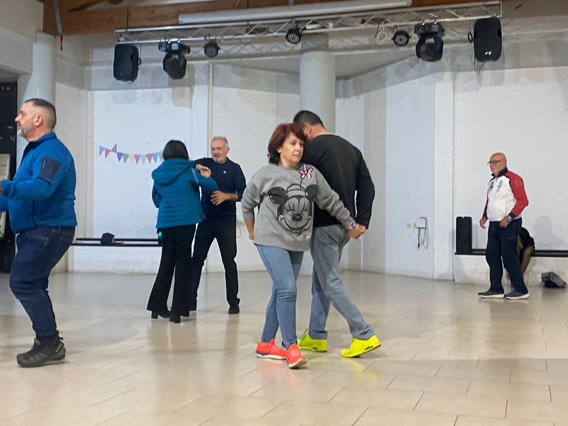Persone che ballano in una sala da ballo illuminata. Alcuni indossano abiti casual. Un palco e delle luci sullo sfondo.