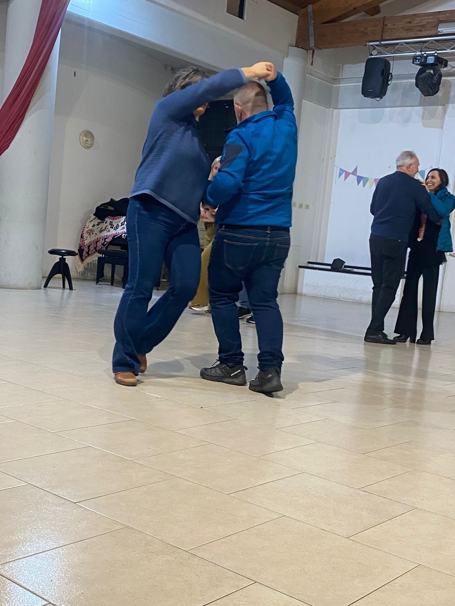 Due uomini ballano in una stanza ben illuminata. Uno conduce il ballo con il braccio alzato, entrambi in jeans e giacca. Un'altra coppia balla lì vicino.