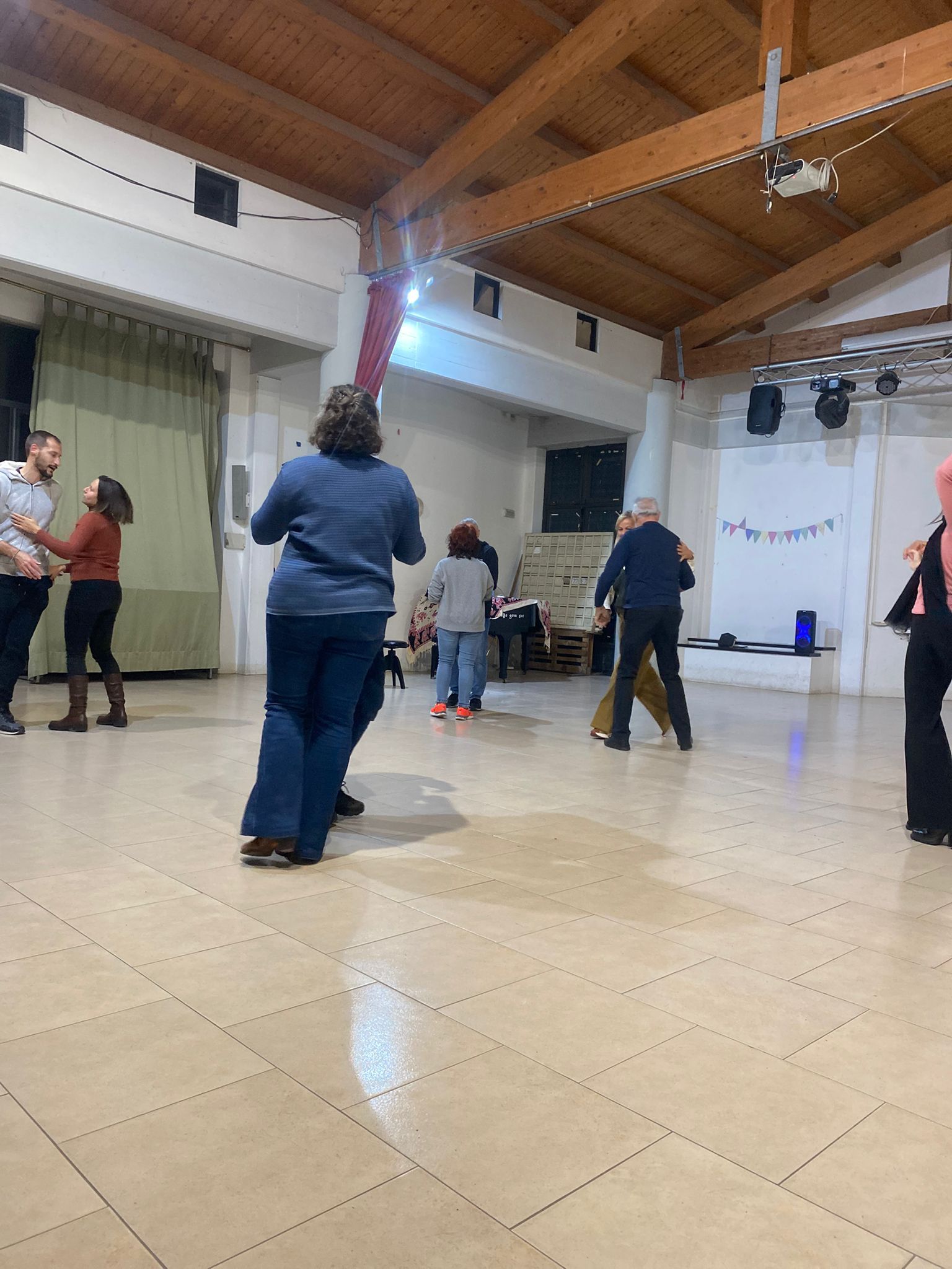 Persone che ballano in una spaziosa sala con travi in ​​legno e pavimento piastrellato.
