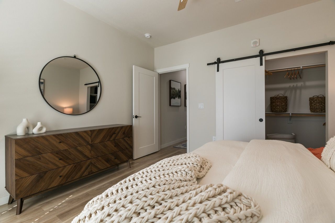 Bedroom with a wooden dresser, round wall mirror, and open closet in neutral tones.