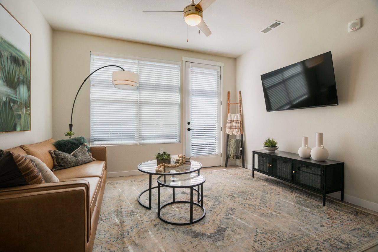Living room in an apartment with a tan sofa, glass coffee tables, a patterned rug, and a wall-mounted TV.