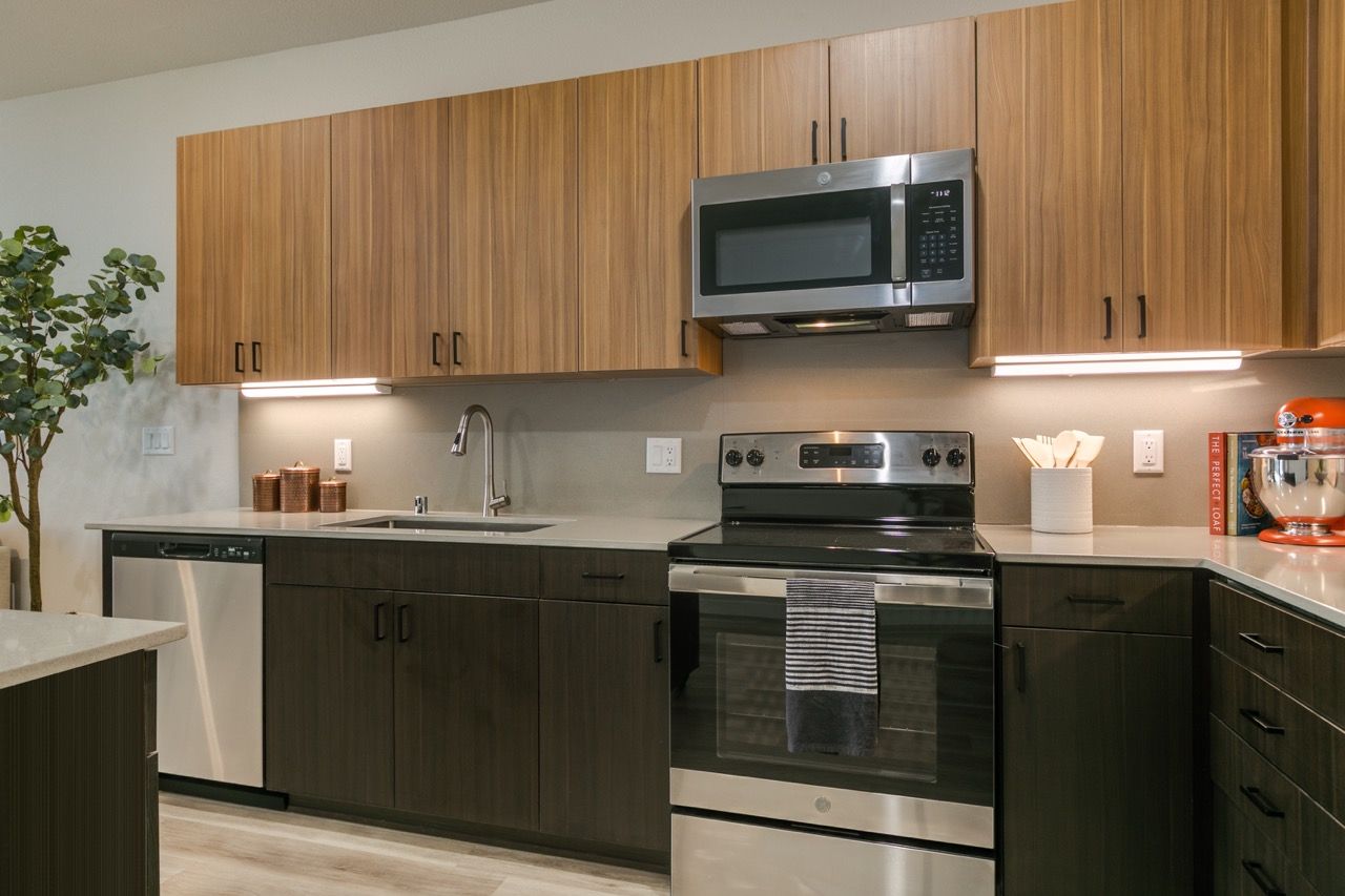 Modern apartment kitchen with wood upper cabinets, dark lower cabinets, stainless steel oven, microwave, and sink.