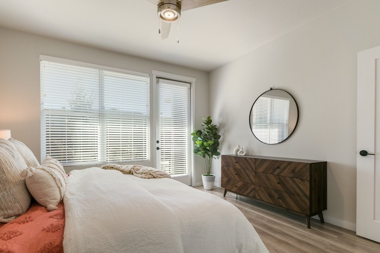 Bedroom in an apartment with a bed, dresser, circular mirror, blinds, and a potted plant.