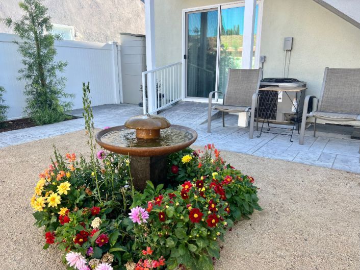 Fountain surrounded by colorful flowers in a backyard with a patio and sliding glass door.