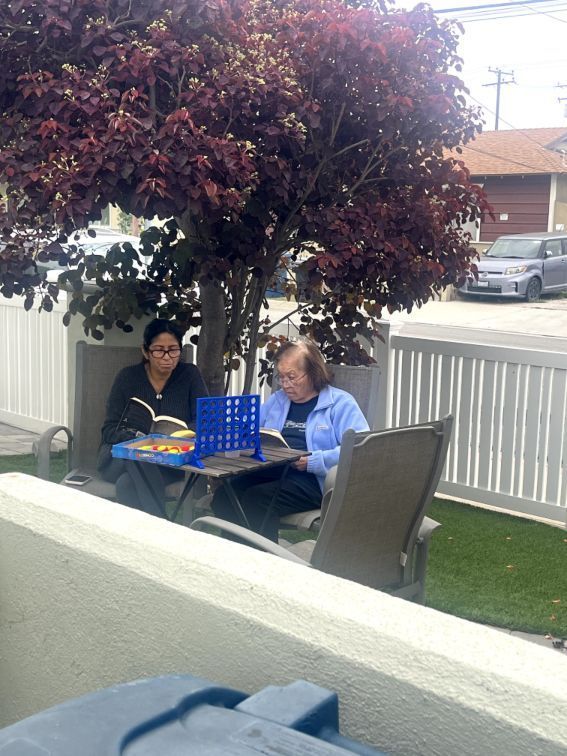 Two women sit outdoors under a tree, playing a board game and reading.. Pepper Tree Assisted Living in Lomita, CA.