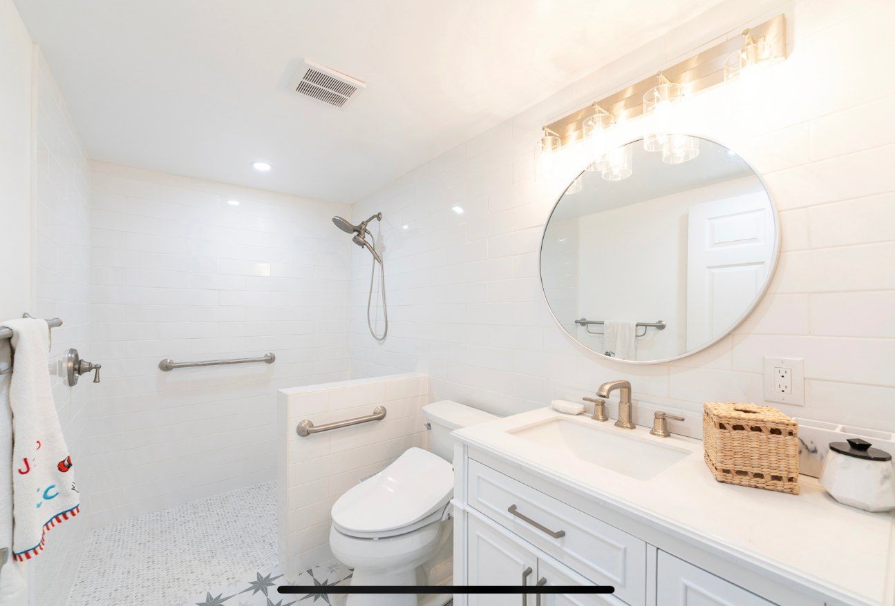 Bright white bathroom with a walk-in shower, toilet, vanity, and round mirror; designed for accessibility.
