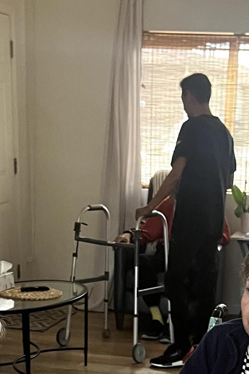 Person assisting someone using a walker indoors; sunny window in background. Pepper Tree Assisted Living in Lomita, CA.