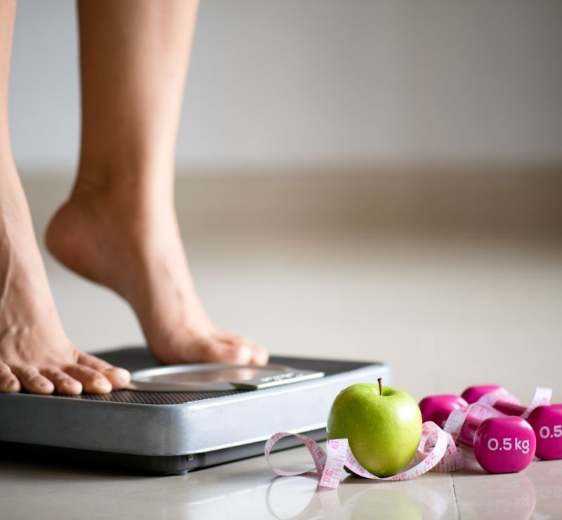 A woman is standing on a scale next to an apple and dumbbells.