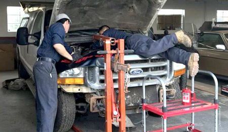 Two mechanics working on a Ford truck engine in a garage. One on the ground, other at the engine bay.