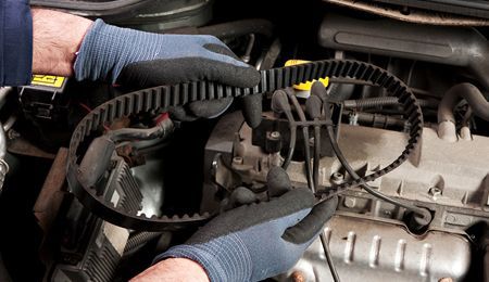 Hands in gloves holding a worn timing belt next to an engine, indicating car maintenance.