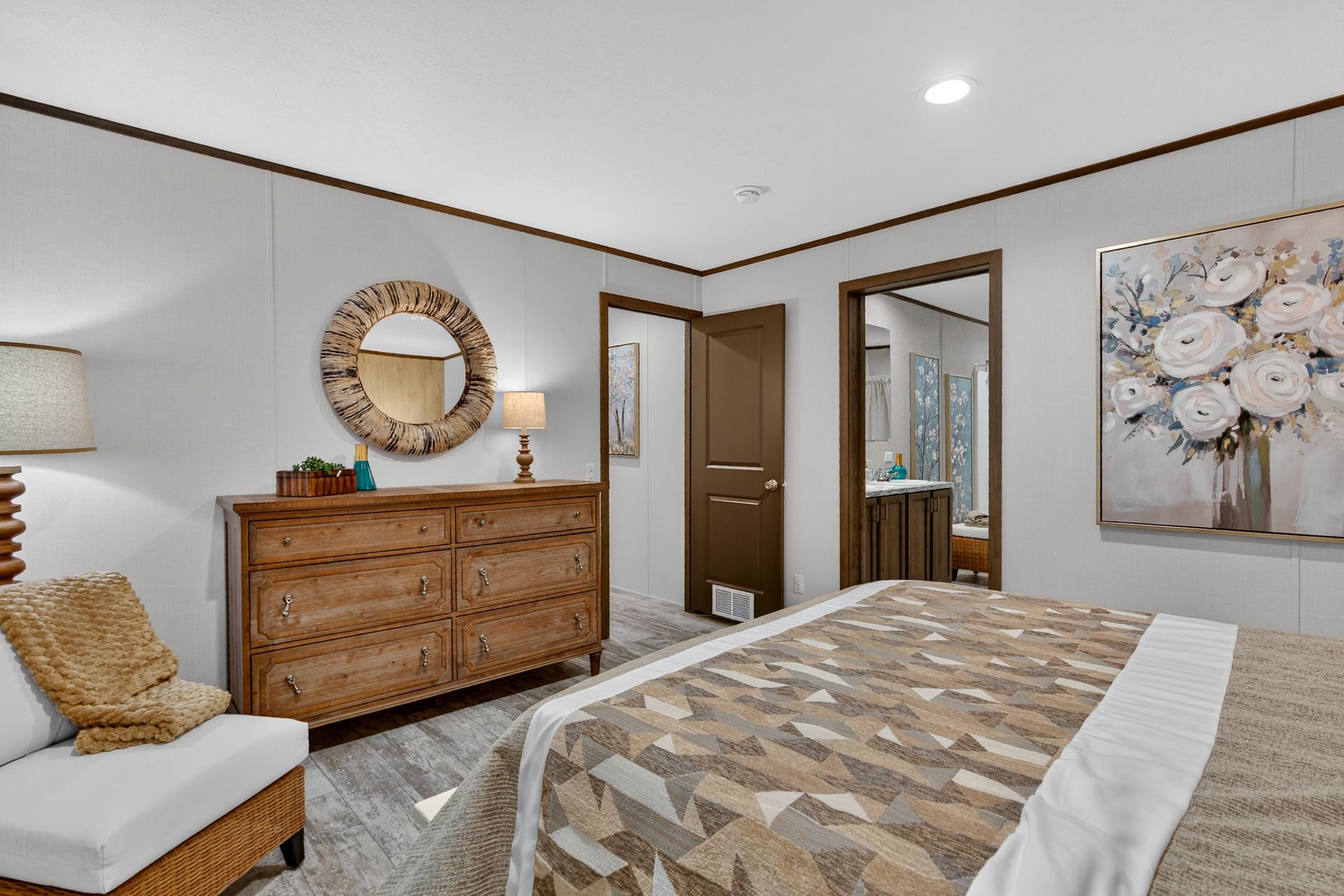 Bedroom with wooden dresser, round mirror, bed, and floral wall art in neutral tones.