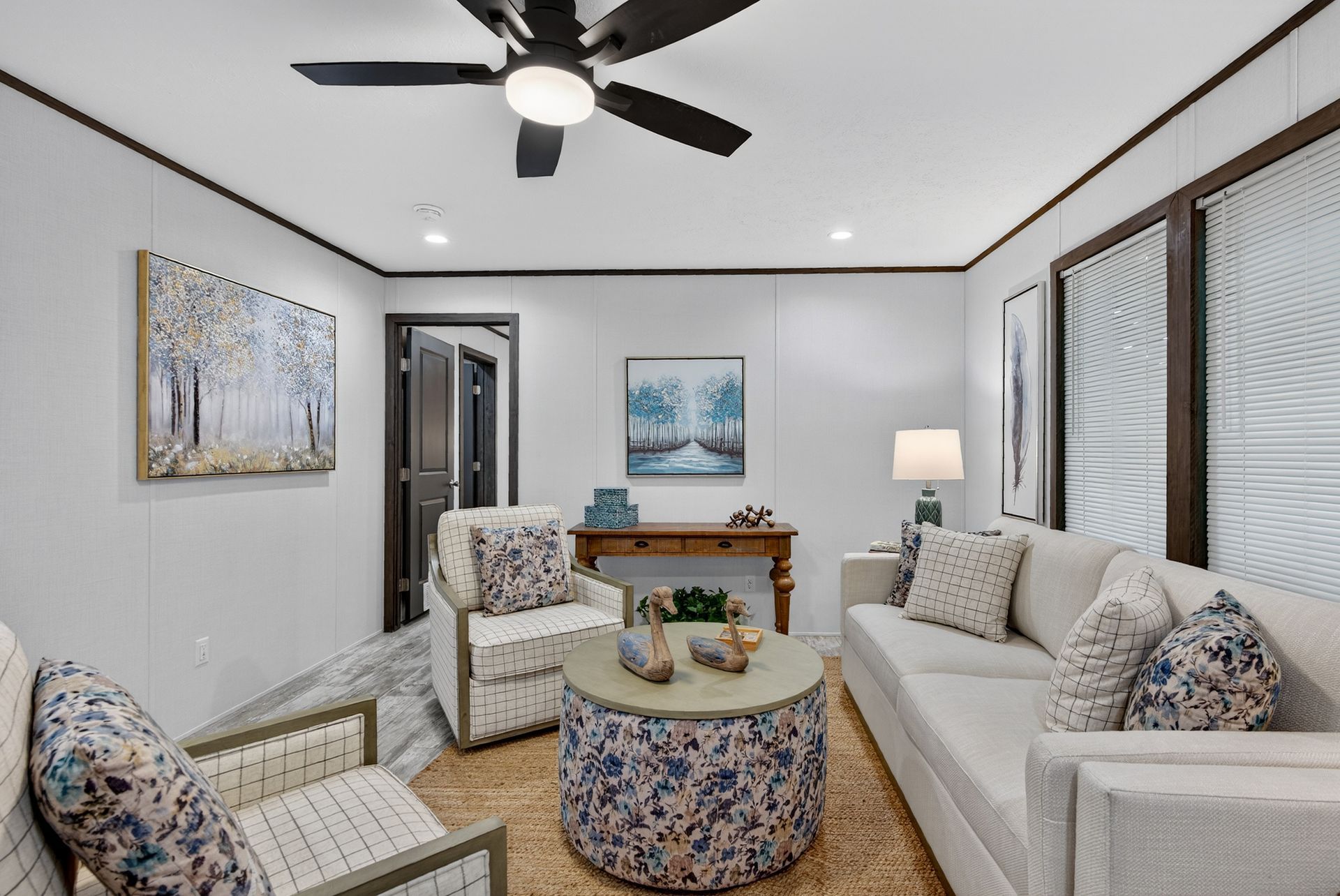 Cozy living room with white sofa, patterned chairs, round ottoman, ceiling fan, and blue artwork