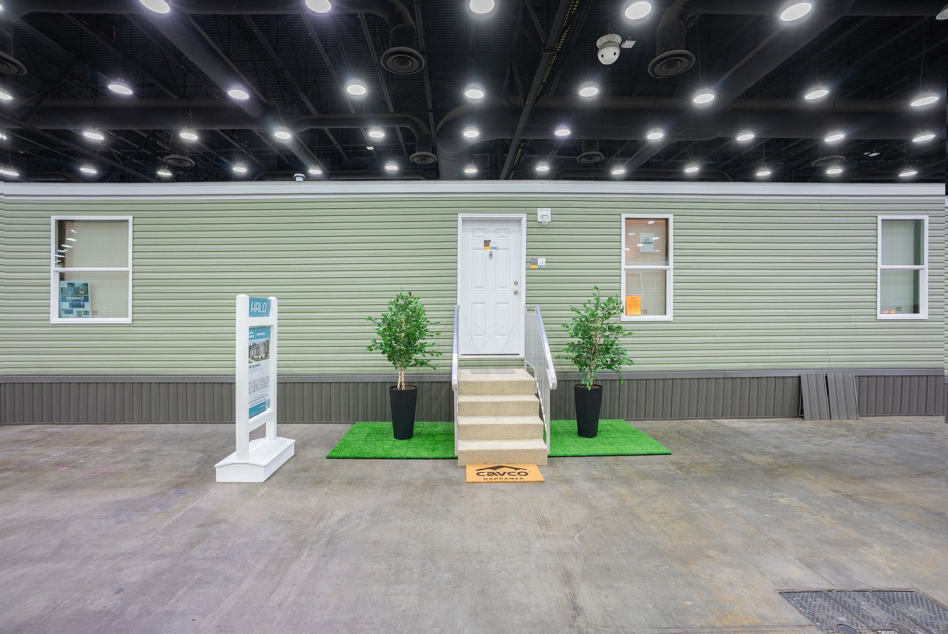 Small green modular home display with white door, steps, and potted plants inside an exhibition hall.
