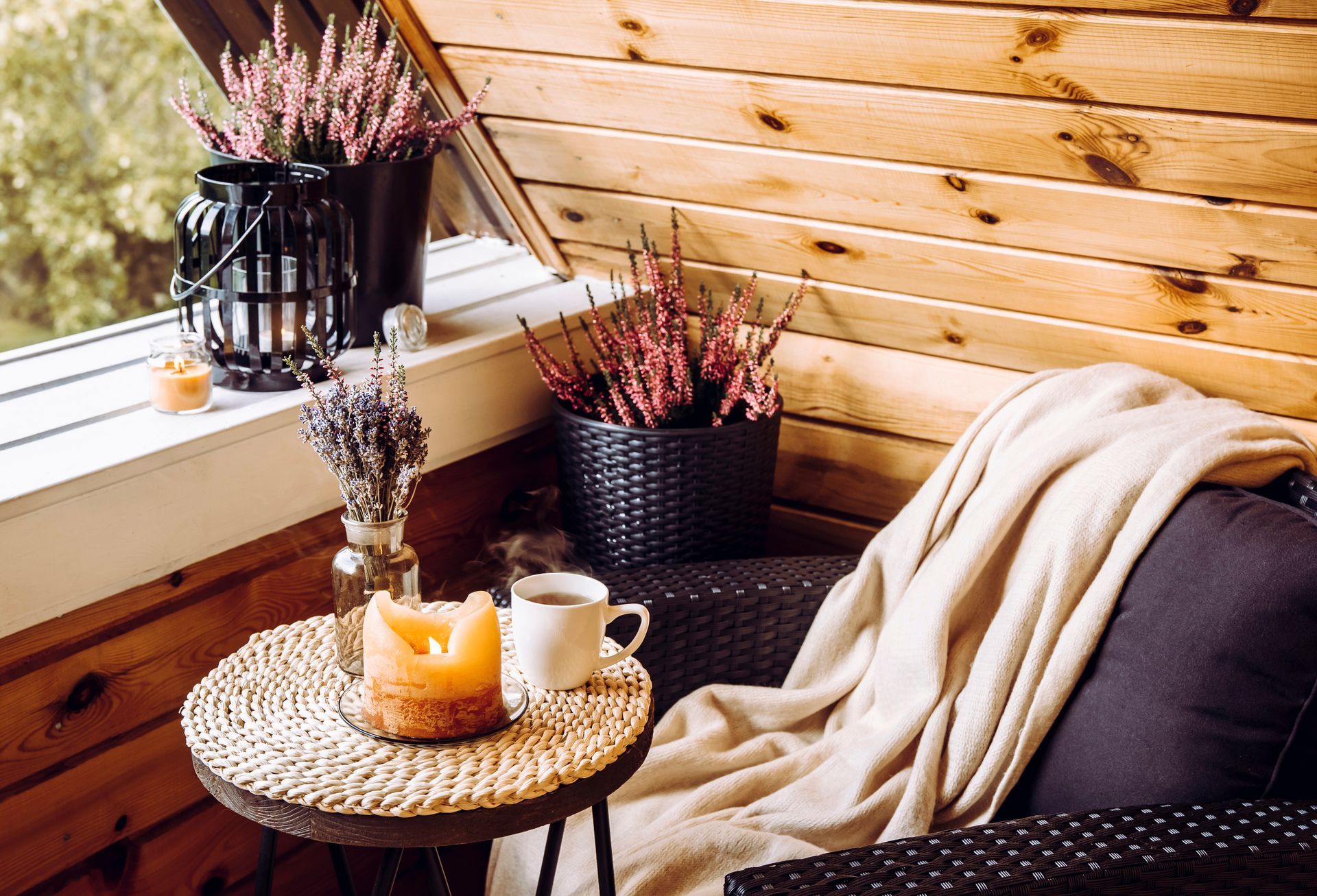 Cozy window seat with woven table, candles, plants, and a cream blanket against a wooden wall