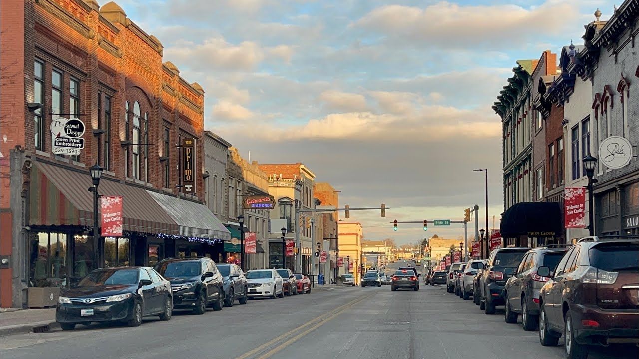 Downtown street lined with brick storefronts and parked cars under a partly cloudy sky