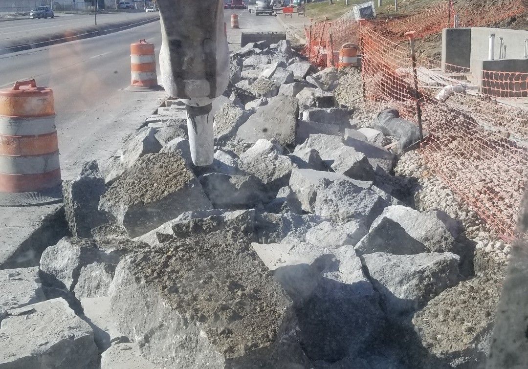 Construction site with jackhammer breaking concrete barrier; orange and white traffic cones, safety fence.