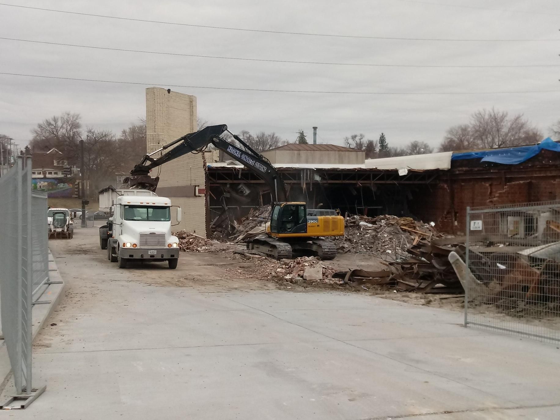 Demolition of a building. Excavator tearing down the structure, a truck waits to haul debris, cloudy sky.