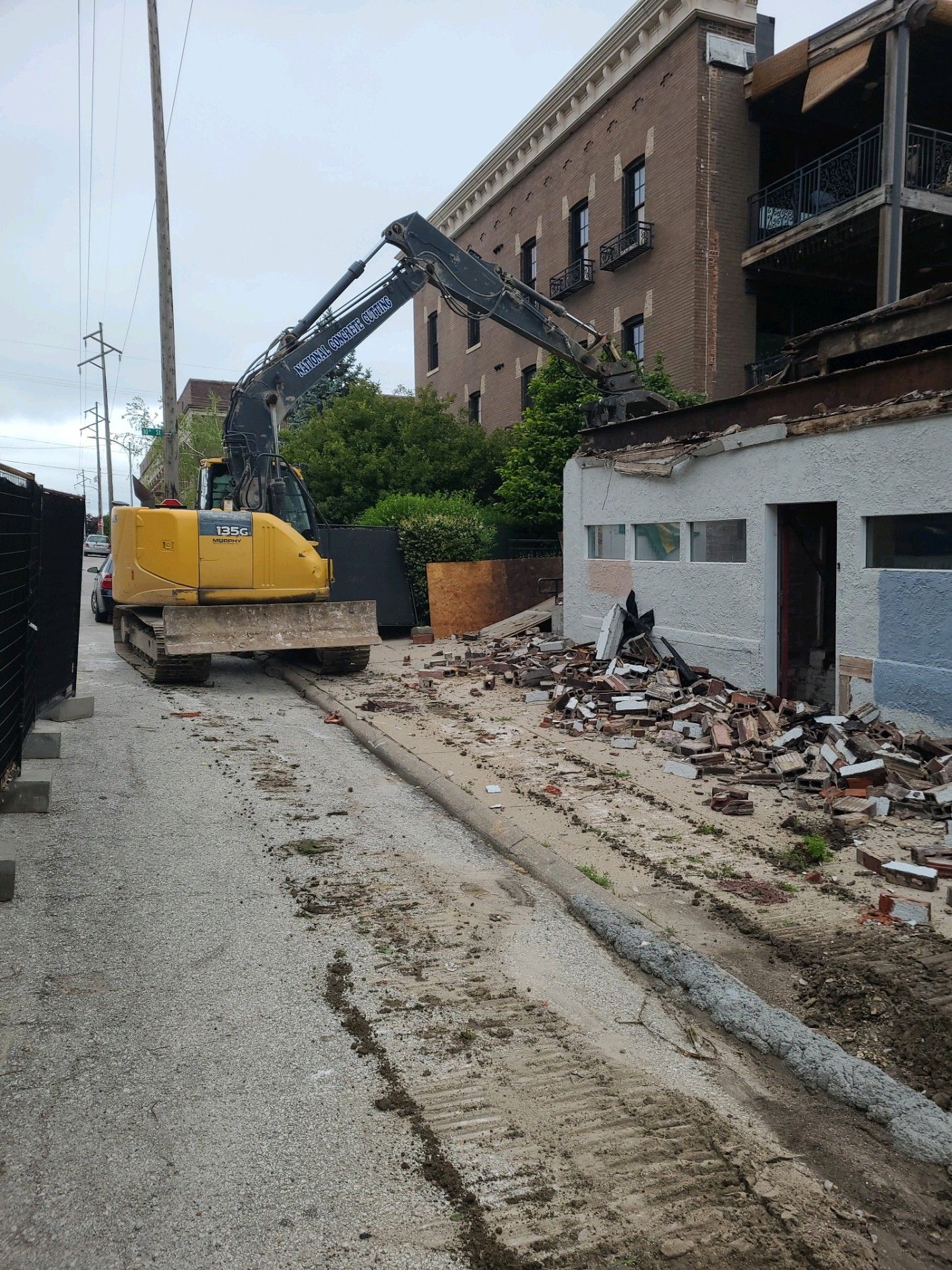 Yellow excavator demolishing a small building next to a brick apartment building. Demolition debris is visible.