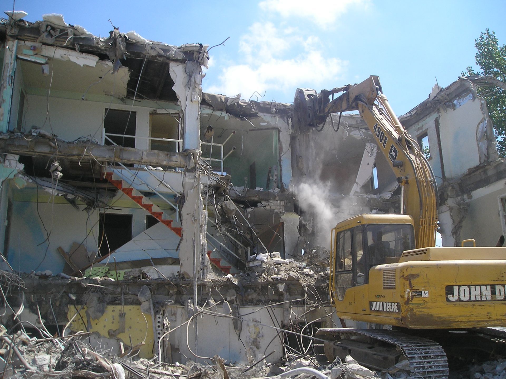 A building being demolished by a yellow excavator on a sunny day. Dust rises as the machine tears down the structure.
