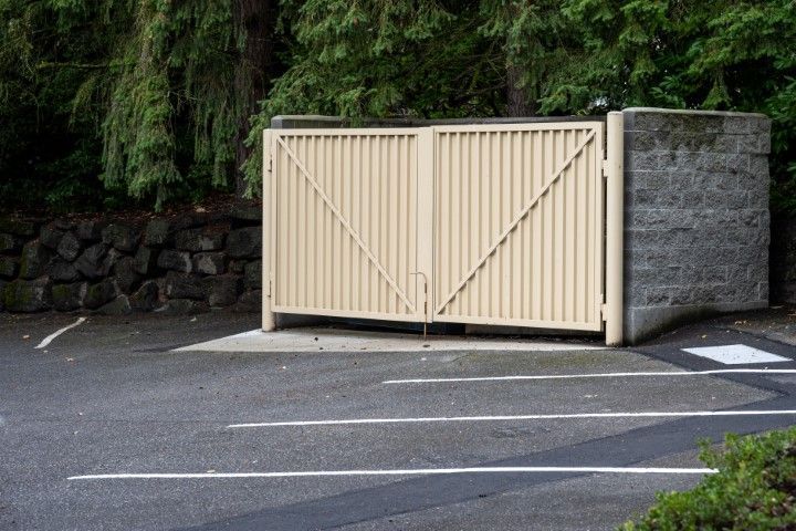 Closed beige metal gate set in a gray stone wall, on a blacktop parking area.