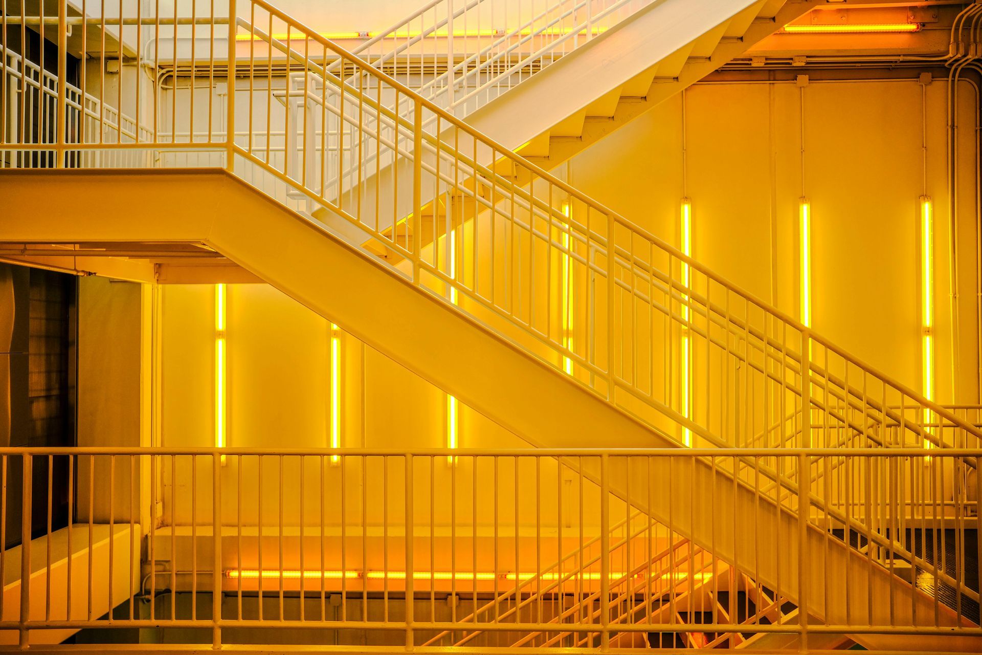 A modern metal staircase bathed in vibrant yellow light, with vertical glowing light fixtures on the walls.