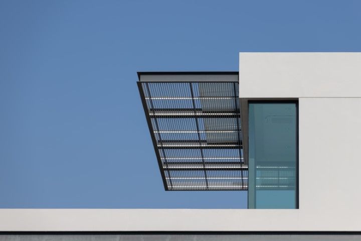 Modern white building corner with a shaded roof and a large window against a blue sky.