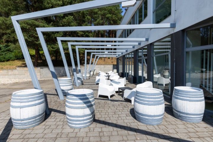 Outdoor seating area with white chairs, gray barrels, and metal overhead structure.
