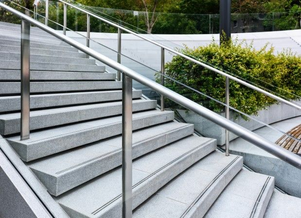 A wide staircase made of gray stone with stainless steel handrails, flanked by green bushes on the right