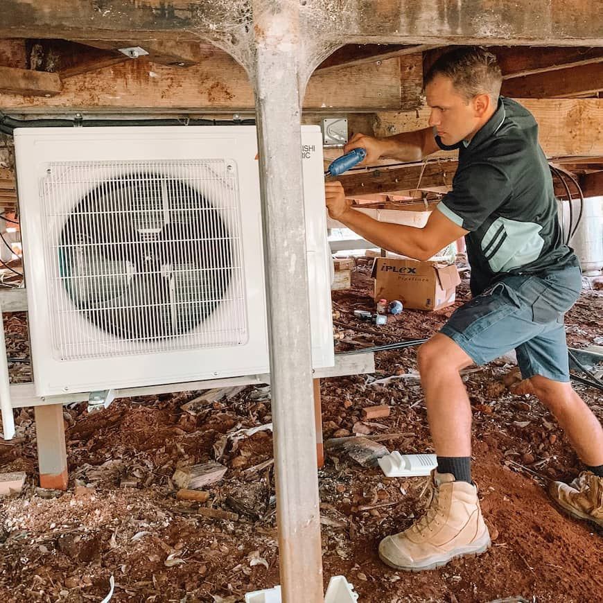 A Man is Working on an Air Conditioner Under a House — Davies Refrigeration & Air Conditioning in Warana, QLD