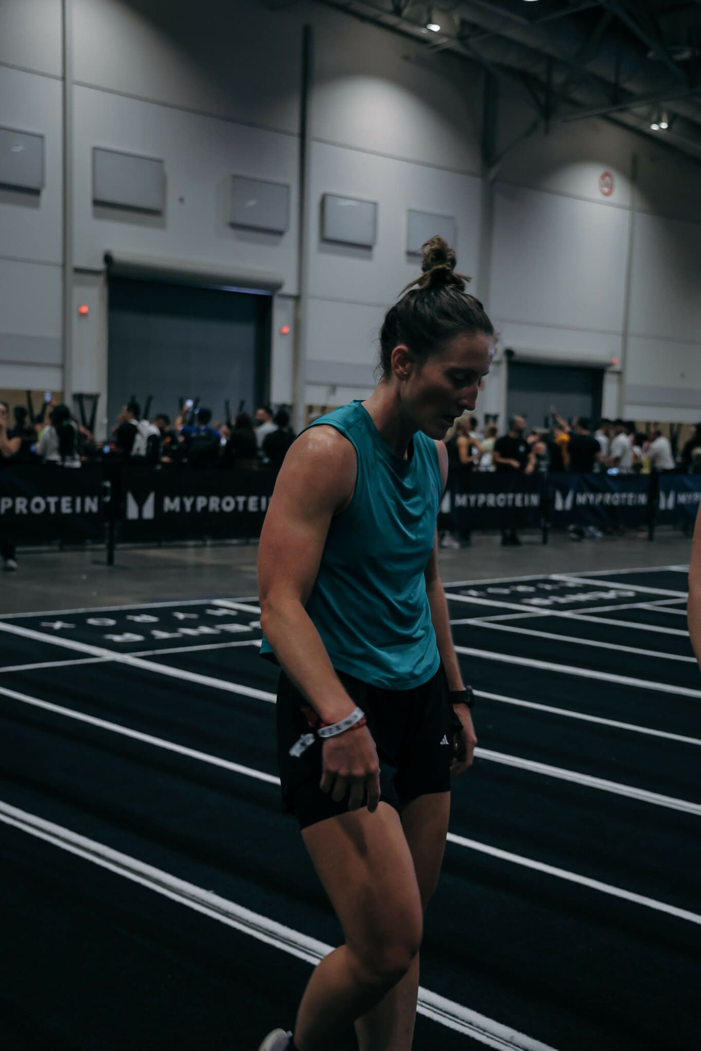 Woman in teal top and black shorts on a track smiles, possibly after a race, with crowd in background.