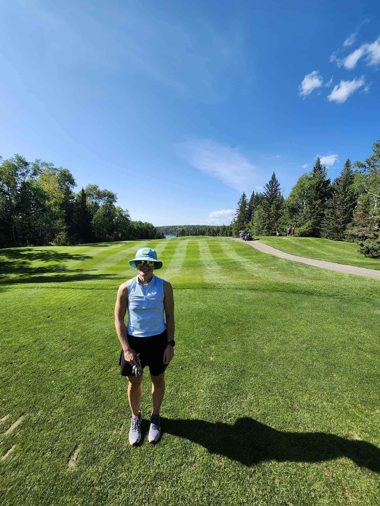 Person stands on a green golf course, blue sky with trees in the background.